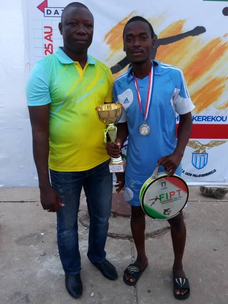 Bénin, juillet 2018 : au Tournoi national du jeu de balle au tambourin, Narcisse Amoussou, en maillot bleu ciel, médaille au cou, tient la coupe du deuxième prix du championnat national, remportée avec l’AS ACADEMIA de Cotonou, finaliste face aux Aigles rouges de Comé, et son tambourin ; à sa droite, en polo jaune et vert, Thomas Tossou-Dan, président de la Fédération béninoise