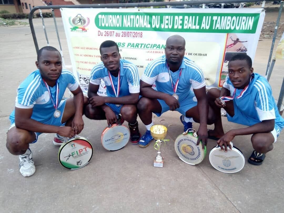 Bénin, juillet 2018 : quatre joueurs de l’AS ACADEMIA de Cotonou, dont Narcisse Amoussou, accroupis en maillot bleu ciel, médaille au cou, autour de la coupe du deuxième prix et de leurs tambourins, devant la banderole du Tournoi national du jeu de balle au tambourin, du 26 au 28 juillet 2018