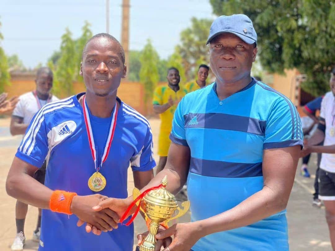 Parakou, 2021, premier tournoi organisé dans le Nord du Bénin : Narcisse Amoussou, de l’équipe des encadreurs, en maillot bleu, médaille d’or au cou, reçoit la coupe, d’une poignée de main, de Thomas Tossou-Dan, président de la Fédération béninoise, en casquette
