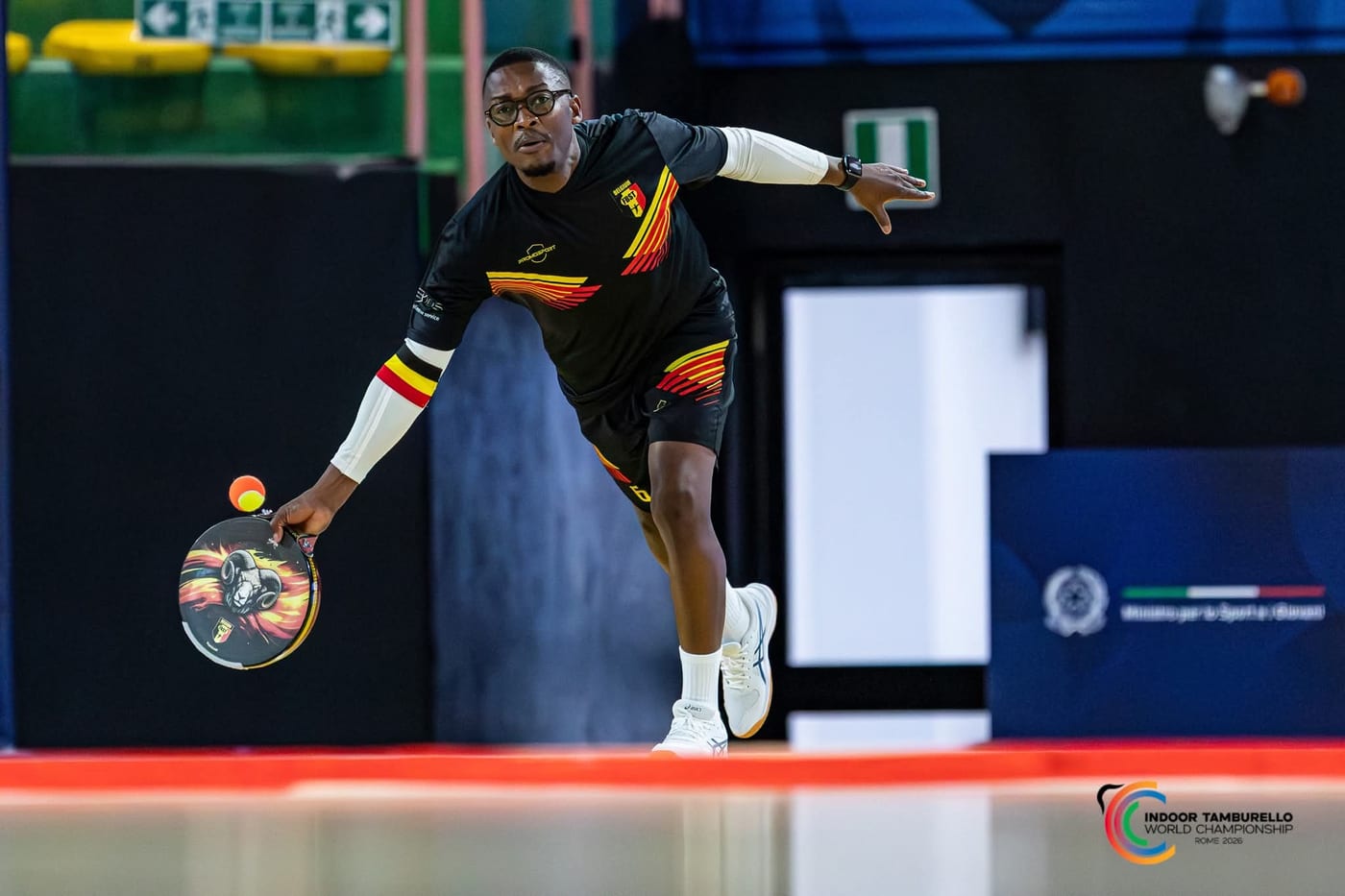 Narcisse Amoussou, en maillot noir, frappe la balle au ras du sol avec son tambourin orné d’un bélier, au Championnat du Monde Indoor de Rome, en 2026